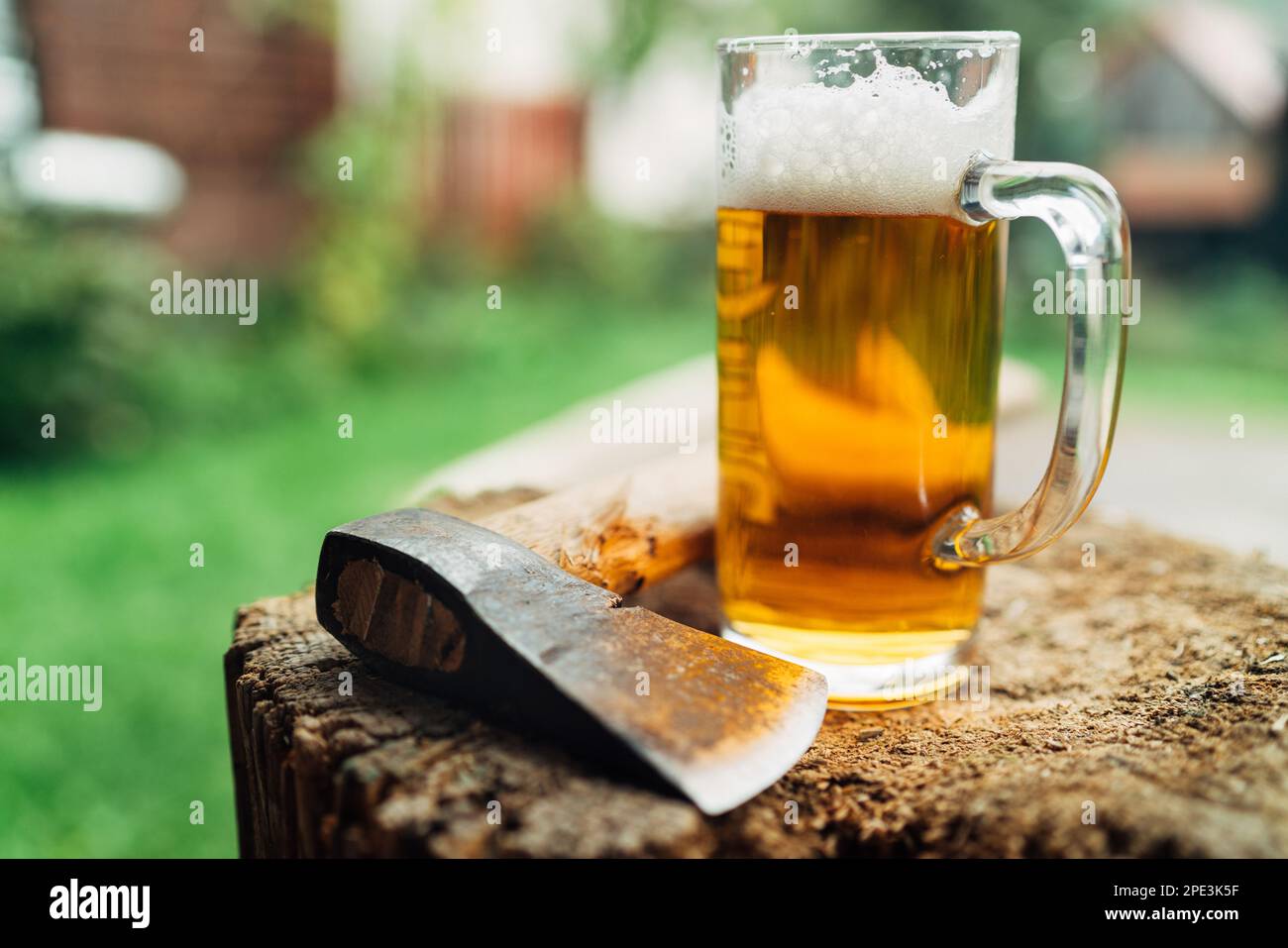 Standbild eines Glases frischen kalten Bieres auf einem Holzstummel neben der Axt. Ländliches Konzept, Männlichkeit, grobes, raues Lifestyle-Konzept in der Wildnis. Stockfoto