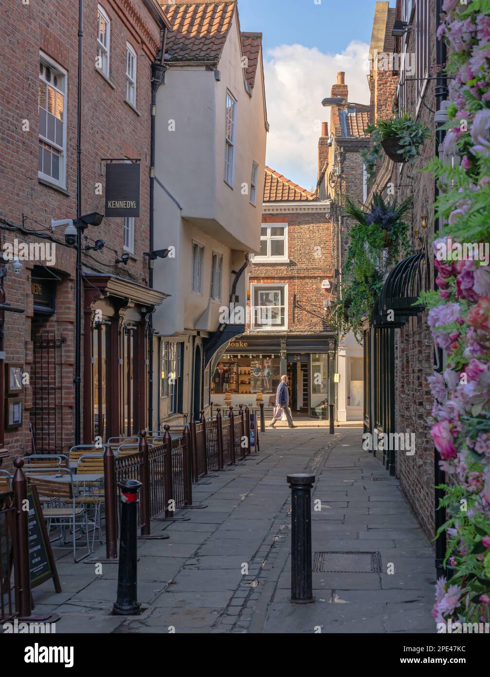 Blick aus einer Gasse auf eine sonnenbeleuchtete Straße. Es gibt ein historisches Gebäude an der Ecke und Tische und Stühle auf einer Seite. Blumen sind im Vordergrund Stockfoto