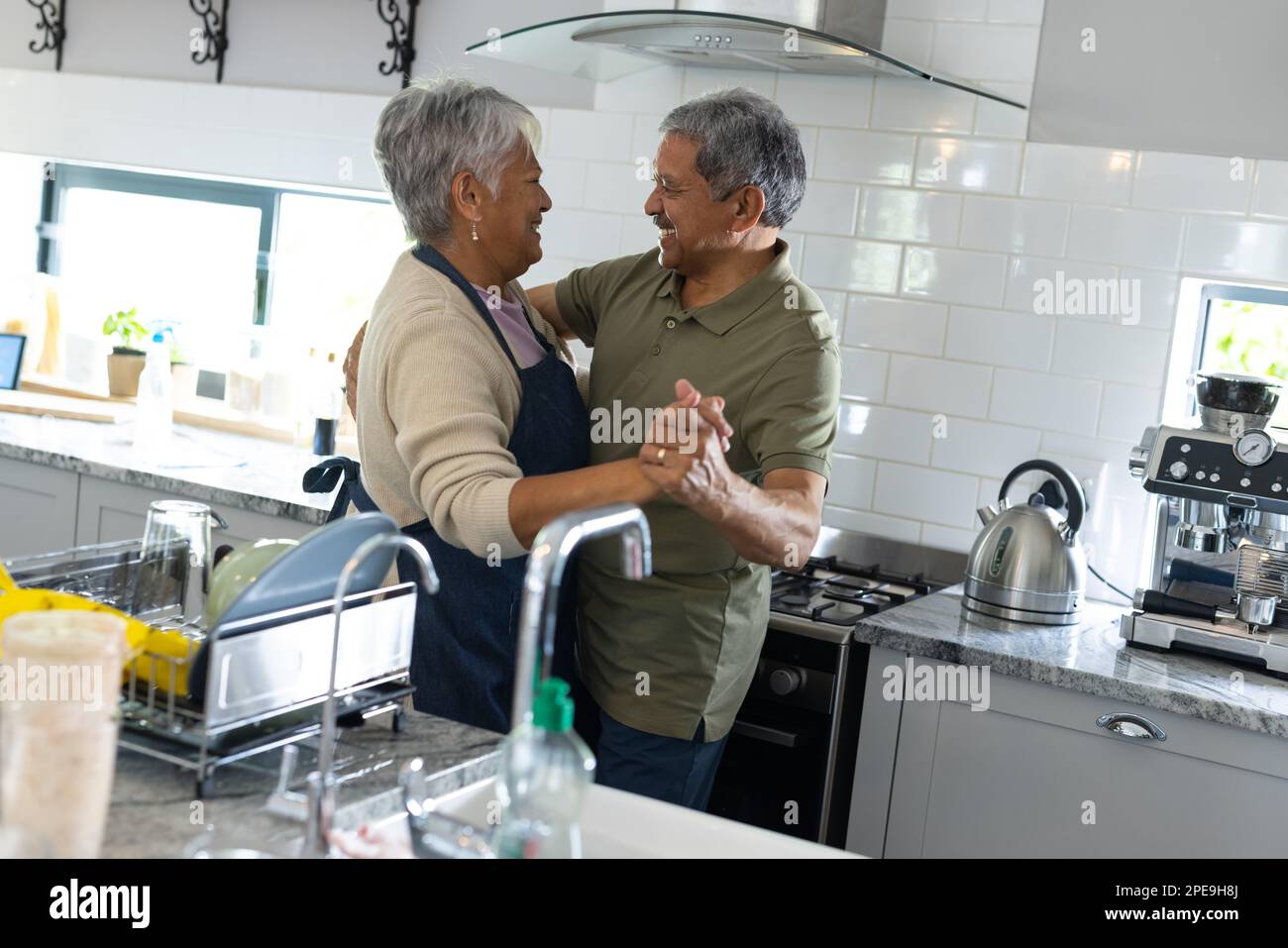 Ein birassistisches Seniorenpaar, das Händchen hält und sich anschaut, während es in der Küche tanzt. Kopierraum, unverändert, Liebe, zusammen, Rentner Stockfoto