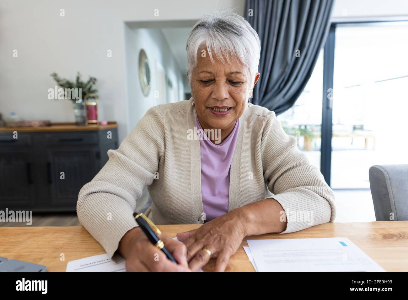 Porträt einer birassischen Seniorin, die Rechnungen auf einem Holztisch analysiert, während sie zu Hause sitzt. Kopierraum, unverändert, Lifestyle, Haushaltsfinanzierung, Budget und RE Stockfoto