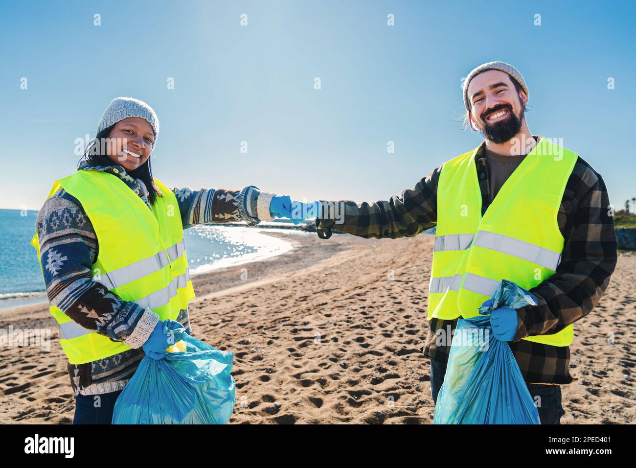 Zwei multiethnische Freiwillige, die Fäuste schlagen, nachdem sie Müll und Plastikschmutz aus dem Sand am Strand aufgesammelt haben. Ein paar Umweltschützer, die lächelnd stehen und in die Kamera schauen. Ökologiekonzept. Hochwertiges Foto Stockfoto