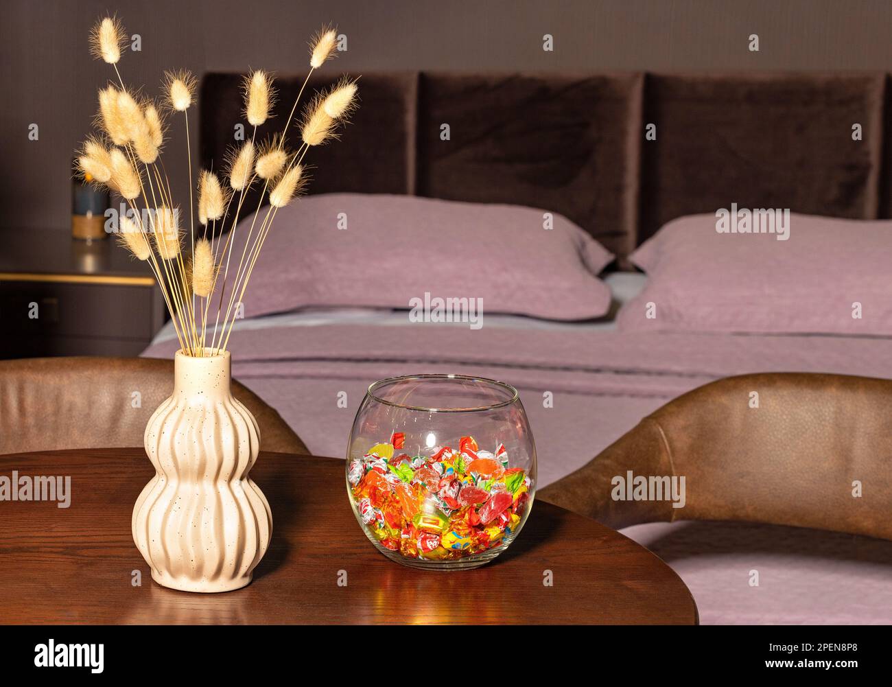 Eine Figur-Steinware-Vase mit flauschigen Grashalmen und eine Glasvase mit bunten Süßigkeiten in einem verschwommenen Schlafzimmer im Hintergrund. Stockfoto