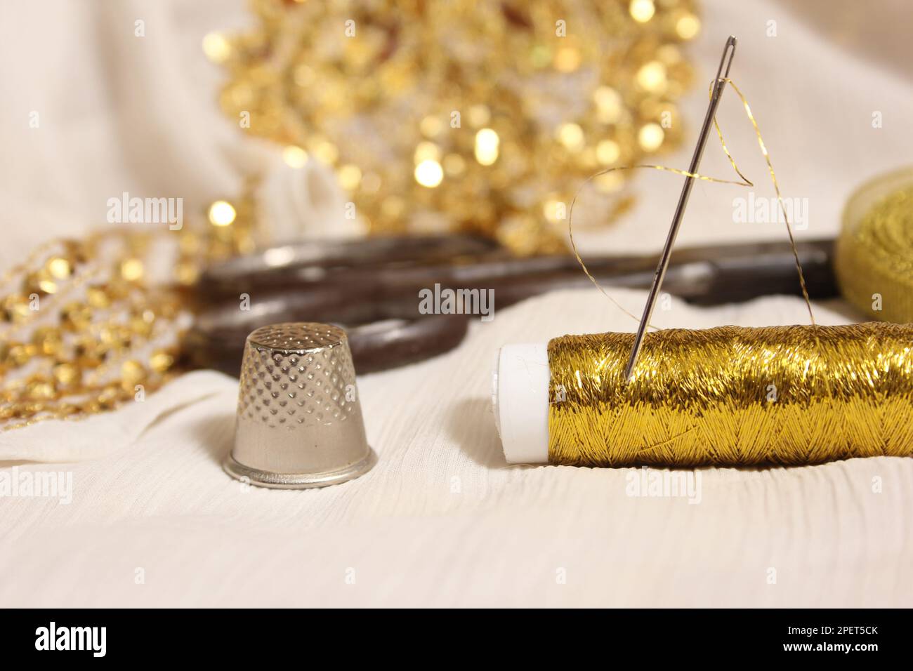 Rolle mit goldmetallischem Gewinde und Schere mit Kaugummi auf weißem Stoff mit goldfarbenen Pailletten Stockfoto