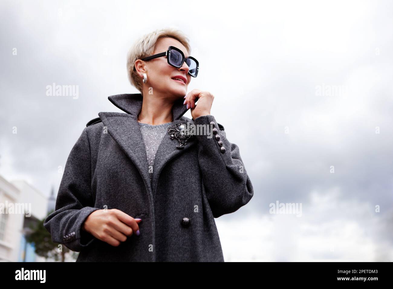 Porträt attraktives weibliches Modell auf bewölktem Himmelshintergrund. Moderne, stilvolle Frau mit kurzen Haaren in grauem, doppelreihigem Erbsenmantel und Brille Stockfoto