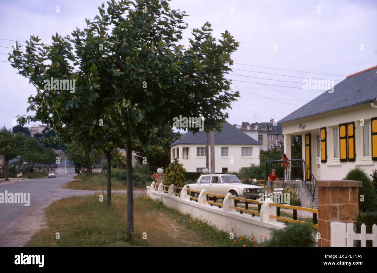 Blick auf die Leve Rames Ferienvilla von der anderen Straßenseite mit einem cremefarbenen Alfa Romeo 2000 Berlina Saloon in der Einfahrt, mit zwei kleinen Kindern auf dem Balkon in St Cast Le Guildo, Bretagne, Frankreich 1971. Stockfoto