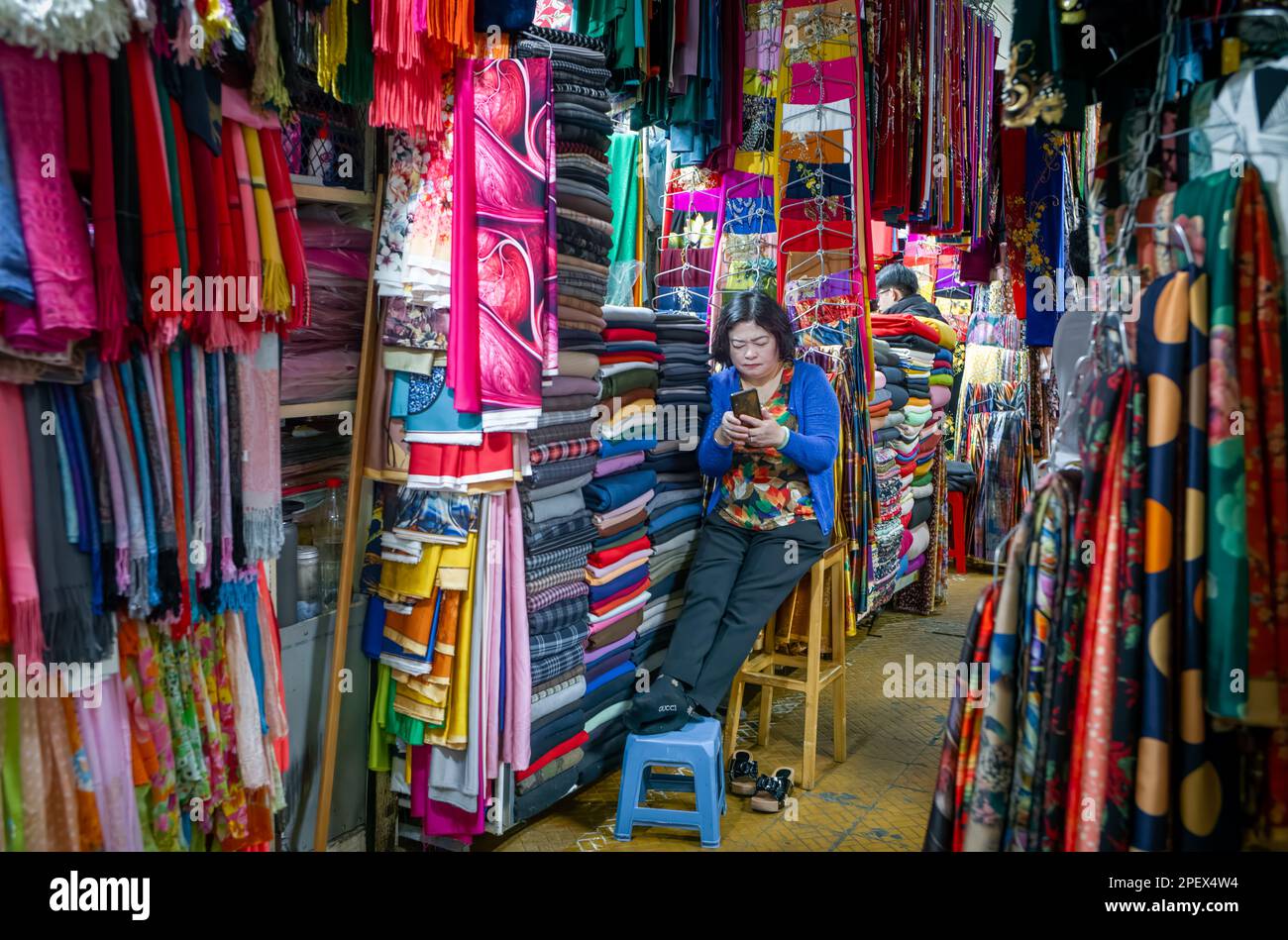 Eine vietnamesische Stallhalterin schaut auf ihr Mobiltelefon, während sie an ihrem Stoffstand im Dalat Market, Vietnam, auf Kunden wartet. Stockfoto