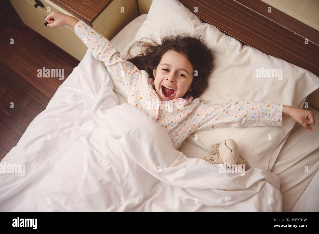 Blick von oben auf das niedliche kleine Mädchen, das die Arme ausstreckt, während es morgens in fröhlicher Stimmung aufwacht Stockfoto