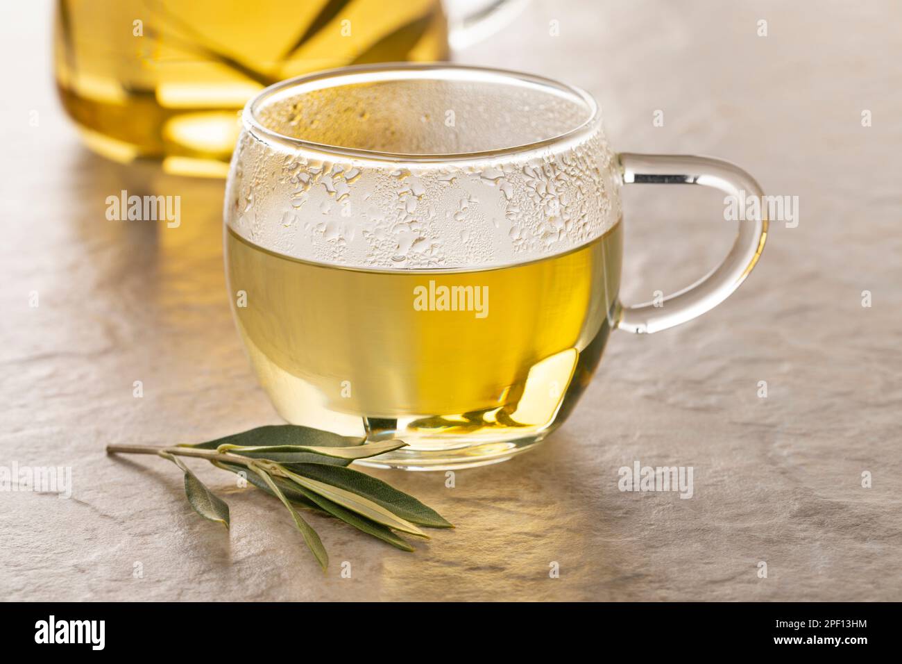 Teeglas mit getrockneten Olivenblättern Tee und einem frischen Zweig Olivenblätter davor Stockfoto