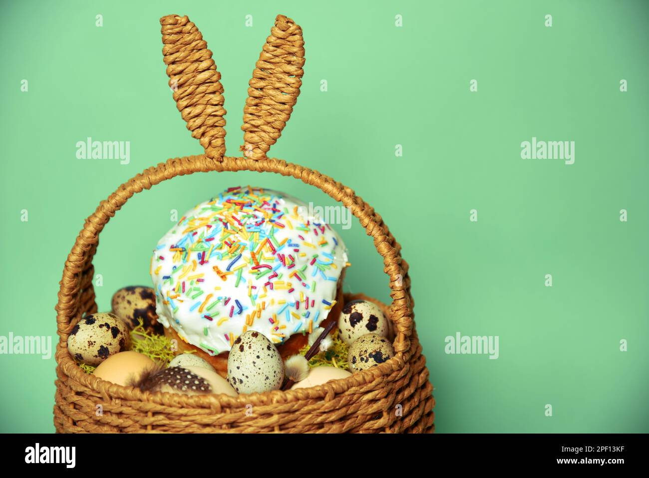 Osterdekorationen in einem Korb mit Kaninchenohren. Osterkuchen und Eier auf Moos, Wachteleier und Federn auf minzgrünem Hintergrund Stockfoto
