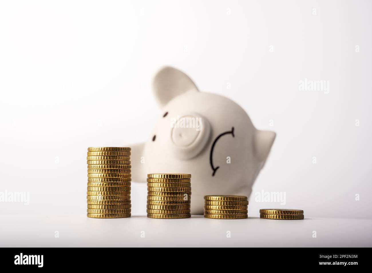 Ein trauriges Sparschwein hinter Euromünzen-Stapeln, das den Fall der Geldwerte symbolisiert. Weißer Geldkasten mit Münzgraph. Krisenkonzept Stockfoto