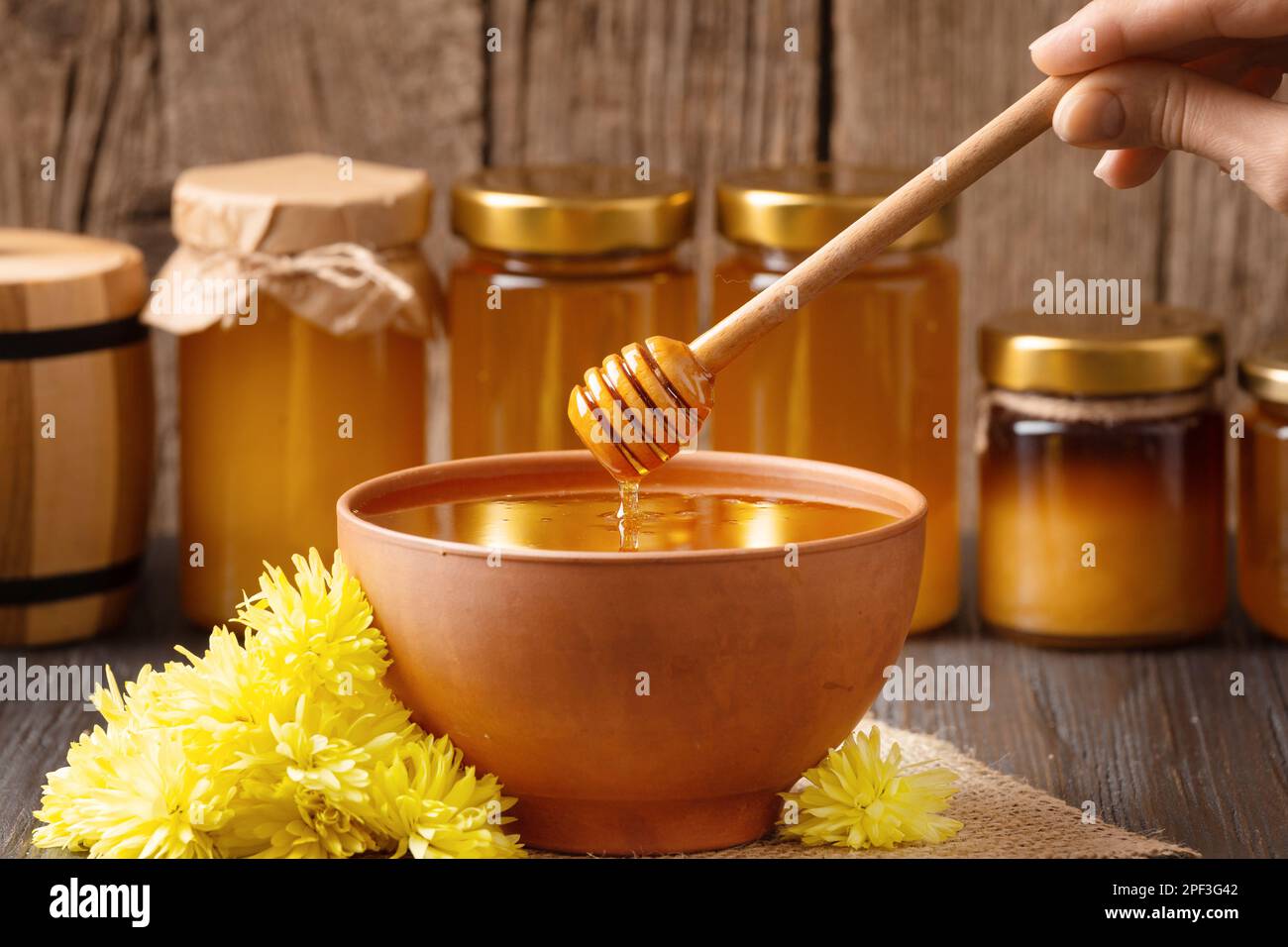 Kreative Zusammensetzung von Gläsern mit Honig, Honig in Tonschüssel, Pendelarm und Honiggießen auf Holzuntergrund. Honig wird aus einem Holzlöffel ausgeschenkt Stockfoto