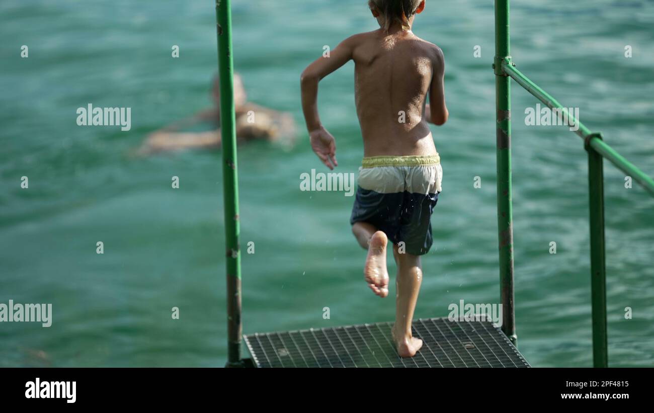Ein Kind, das in das Wasser des Sees stürzt. Das Kind hat Spaß dabei, am Dock vorbeizulaufen und springt in Super Slow Motion in den Fluss Stockfoto