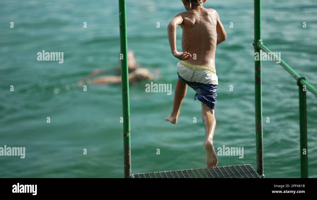 Ein Kind, das in das Wasser des Sees stürzt. Das Kind hat Spaß dabei, am Dock vorbeizulaufen und springt in Super Slow Motion in den Fluss Stockfoto