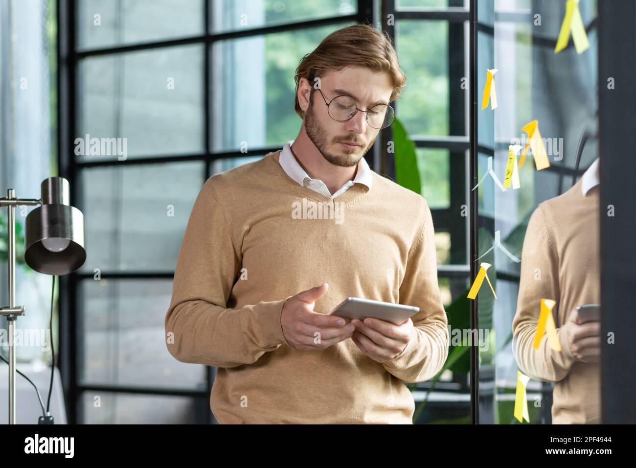 Junger fokussierter männlicher Student, Praktikant im Online-Kurs, der auf den Tablet-Bildschirm schaut, neben einem transparenten Whiteboard mit Notizen steht und sich eine Videokurse ansieht. Stockfoto