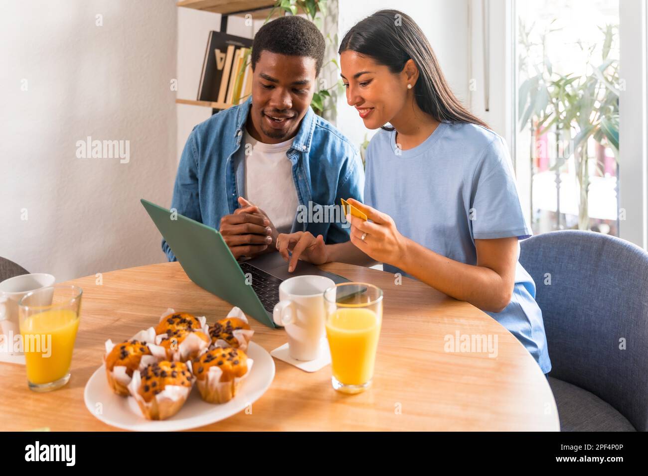Ein multiethnisches Paar kauft online mit dem Computer, während es frühstückt, direkt am Fenster Stockfoto