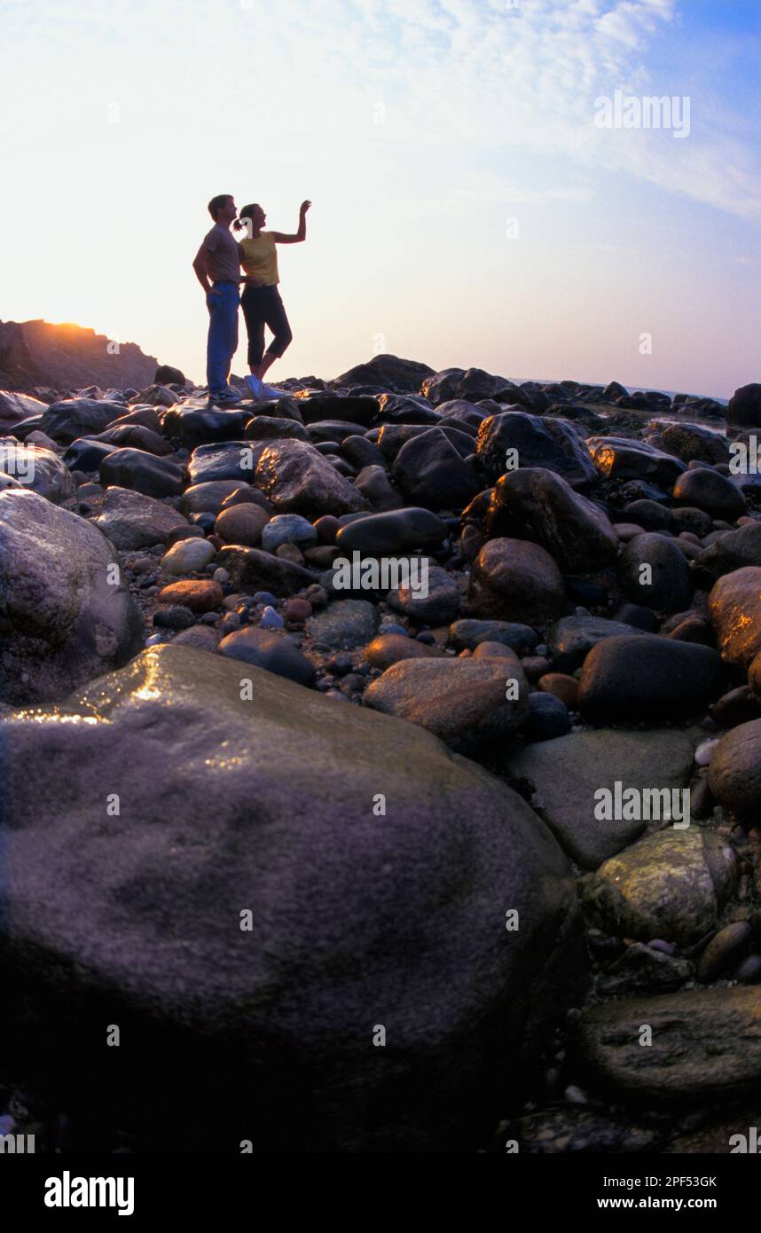 Silhouette eines Paares, das bei Sonnenaufgang an einem felsigen Strand steht Stockfoto