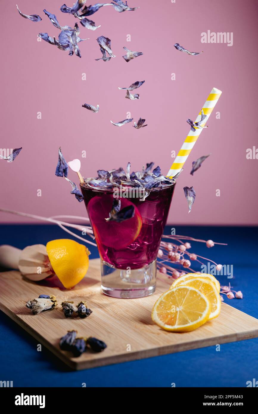 Sommerlich lila Getränk/Cocktail mit Blütenblättern und gelbem Strohhalm, Zitronenscheiben und Eis, mit farbigem Hintergrund Stockfoto