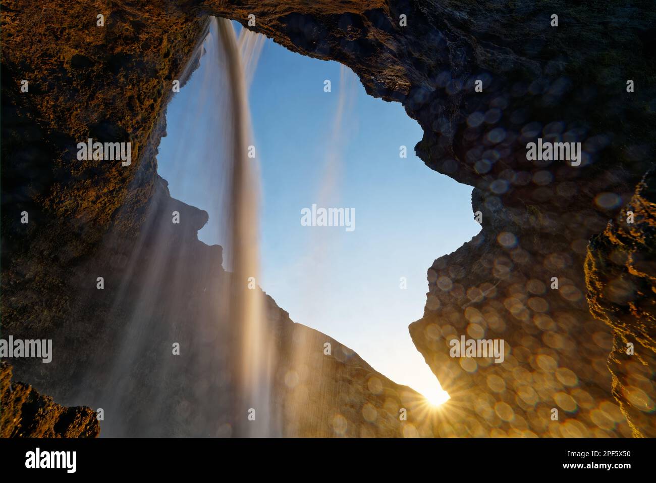 Blick von diagonal unter einem Wasserfall, umgeben von Felsen, die untergehende Sonne mit einem Strahlhalo ist am Rand der Aussichtsplattform zu sehen Stockfoto