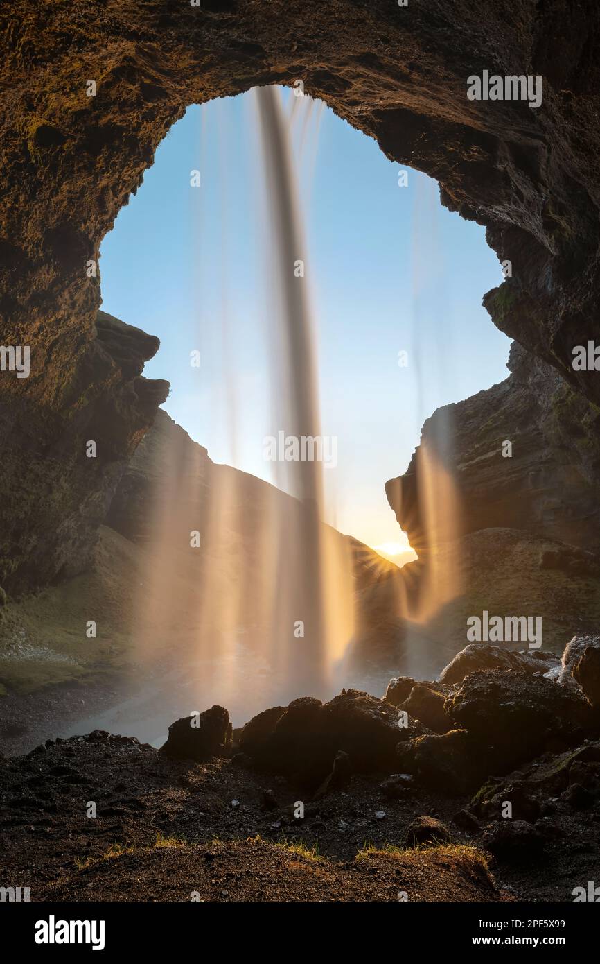 Blick durch einen Wasserfall bei Sonnenuntergang in Island, die untergehende Sonne färbt das Wasser, die Aussicht deckt auch die Gegend vor dem Wasserfall ab, Überreste von Stockfoto