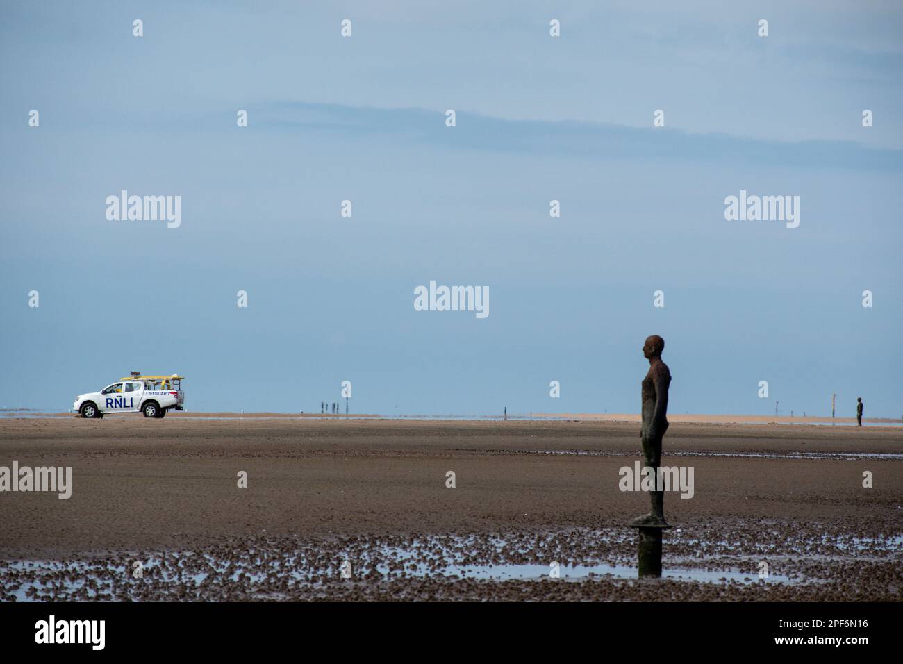 Figur der Installation des Künstlers Antony Gormley an einem anderen Ort am Crosby Beach mit Blick auf das Meer und einem RNLI Life Guard 4x4 im Hintergrund Stockfoto