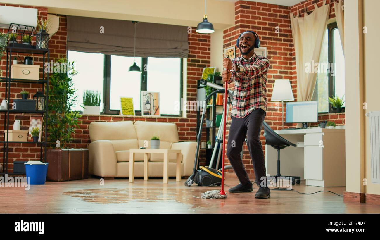 Junger männlicher Erwachsener, der zu Hause Spaß mit Mopp und Musik hat, Tanzbewegungen zeigt und im Wohnzimmer singt. Glückliche Person, die Holzböden gewischt und Frühjahrsputz und Hausarbeit macht. Stockfoto
