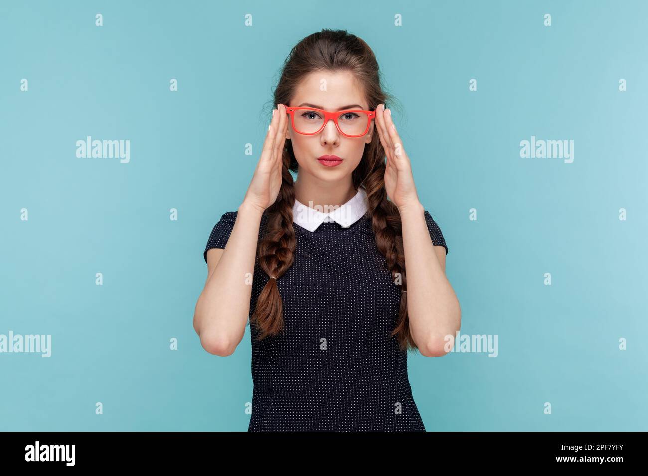 Das Porträt einer attraktiven Frau mit Geflechten hält die Hände auf dem Rahmen einer roten Brille, schaut aufmerksam in die Kamera und trägt ein schwarzes Kleid. Frau Indoor Studio Aufnahme isoliert auf blauem Hintergrund Stockfoto