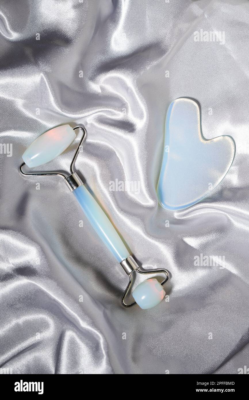 Blaue Jade-Gesichtswalze und Gua Sha-Kratzer für Schönheitsmassagen auf silbernem, strukturiertem Seidenhintergrund. Gesichtsbehandlung und Heim-Spa, Anti-agi Stockfoto