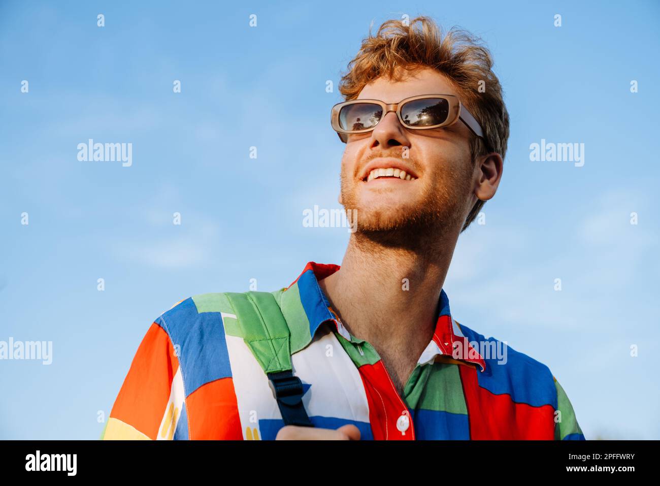 Porträt eines jungen, attraktiven, stylischen, lächelnden Mannes in einem farbenfrohen Hemd und einer Sonnenbrille, die zur Seite blickt Stockfoto
