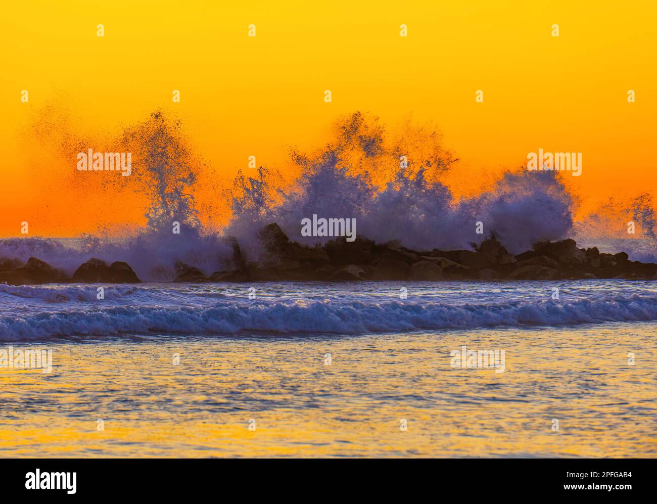 Küstenlandschaft mit dramatischen Wellen, die an der felsigen Küste unter einem farbenfrohen und lebendigen Himmel bei Sonnenuntergang aufschlagen, was einen auffälligen Kontrast zu dem bildet Stockfoto