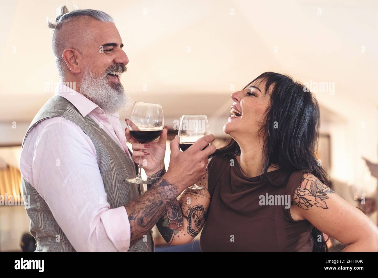 Zwei Hipster-Liebhaber mittleren Alters, ein Mann mit einem langen weißen Bart und Pferdeschwanz, und eine brünette Frau, die in einem verschwommenen Hintergrund steht und auf ihren Sieg anstoßen Stockfoto