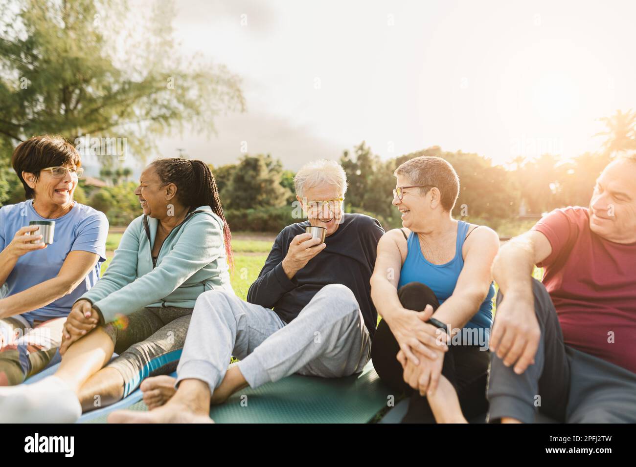 Fröhliche, mehrköpfige Seniorenfreunde, die nach dem Training in einem Park einen Tee trinken Stockfoto