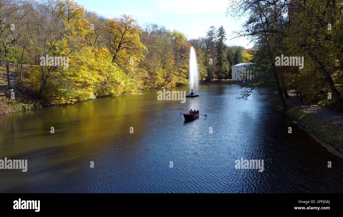 Wunderschöne Landschaften Menschen segeln im Boot auf dem See mit dekorativem Brunnen inmitten von Bäumen mit gelben Blättern im Park an sonnigen Herbsttagen. Natürlicher Hintergrund. Reisetourismus Erholung. Luftaufnahme Stockfoto