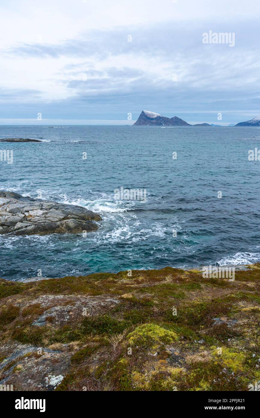 Stürmischer Ozean am Ufer von Hillesøya, in Troms, Norwegen, mit Wellen an der Küste kleiner Inseln, mit schneebedeckten Bergen im Hintergrund. Stockfoto