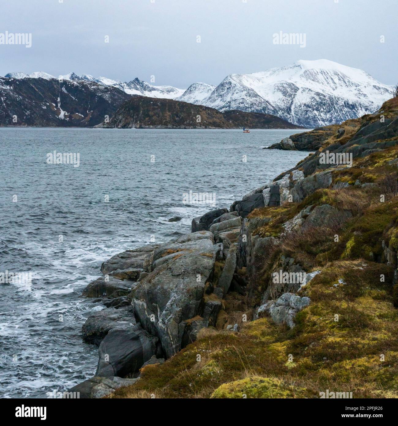 Stürmischer Ozean am Ufer von Hillesøya, in Troms, Norwegen, mit Wellen an der Küste kleiner Inseln, mit schneebedeckten Bergen im Hintergrund. Stockfoto