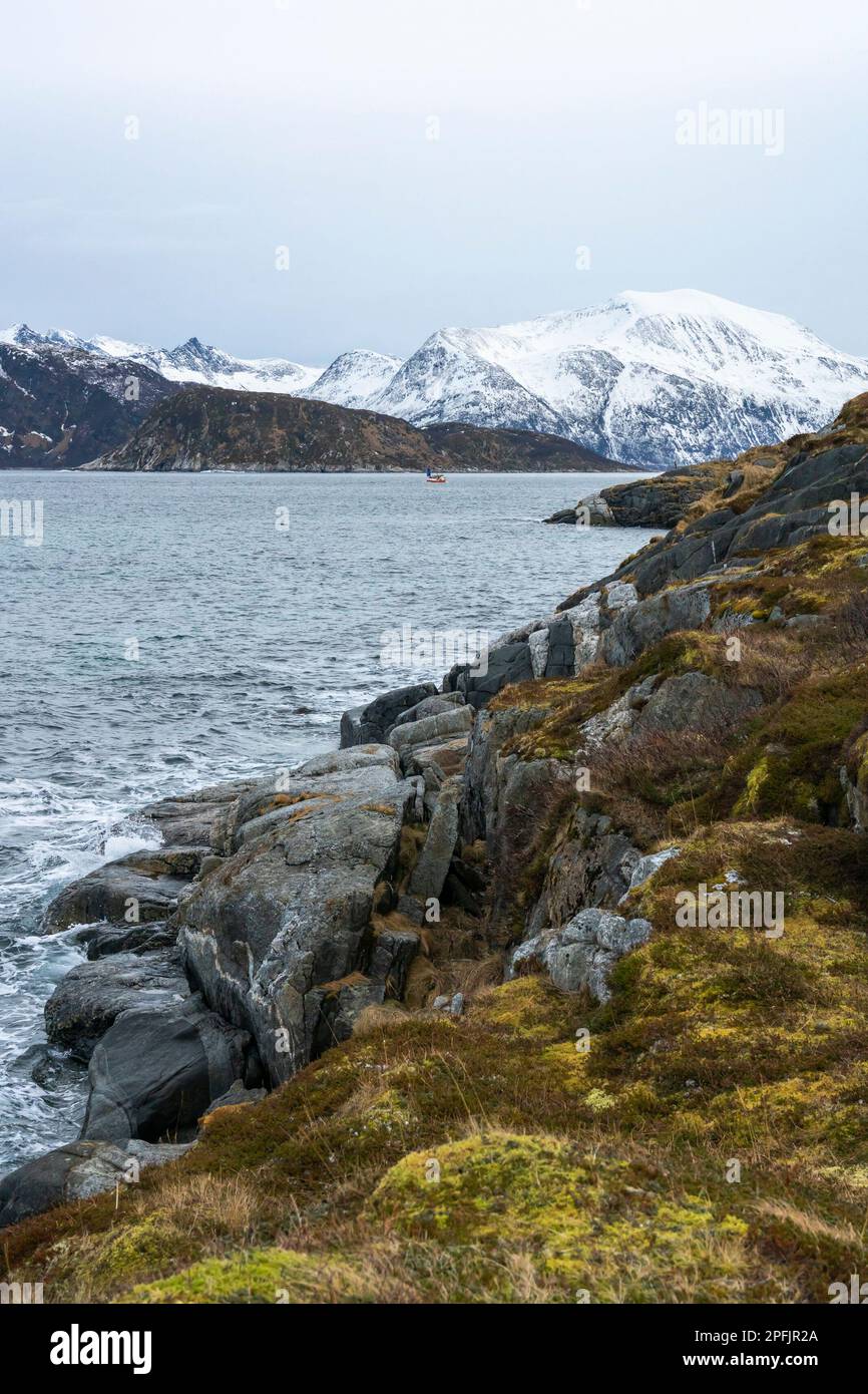 Stürmischer Ozean am Ufer von Hillesøya, in Troms, Norwegen, mit Wellen an der Küste kleiner Inseln, mit schneebedeckten Bergen im Hintergrund. Stockfoto
