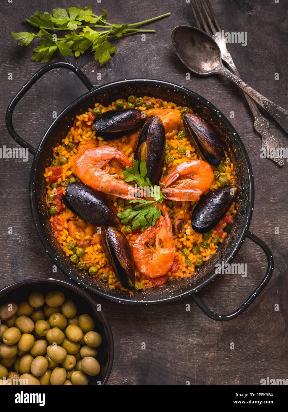 Paella in schwarzer Pfanne mit Reis, Garnelen, Muscheln, Tintenfisch und Fleisch, Schüssel mit Oliven und altem Besteck. Meeresfrüchte-Paella, traditionelles spanisches Gericht. Stockfoto