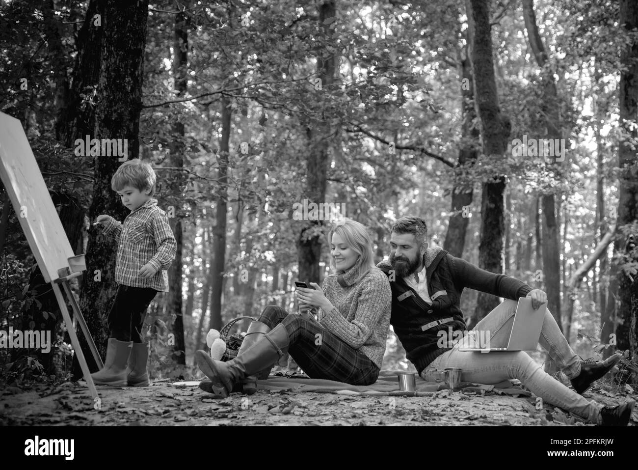 Lächelnde junge Familie, die an einem Herbsttag im Wald in Blättern sitzt. Eine glückliche dreiköpfige Familie verbringt viel Spaß im Park. Der kleine Junge malt mit einem Stockfoto