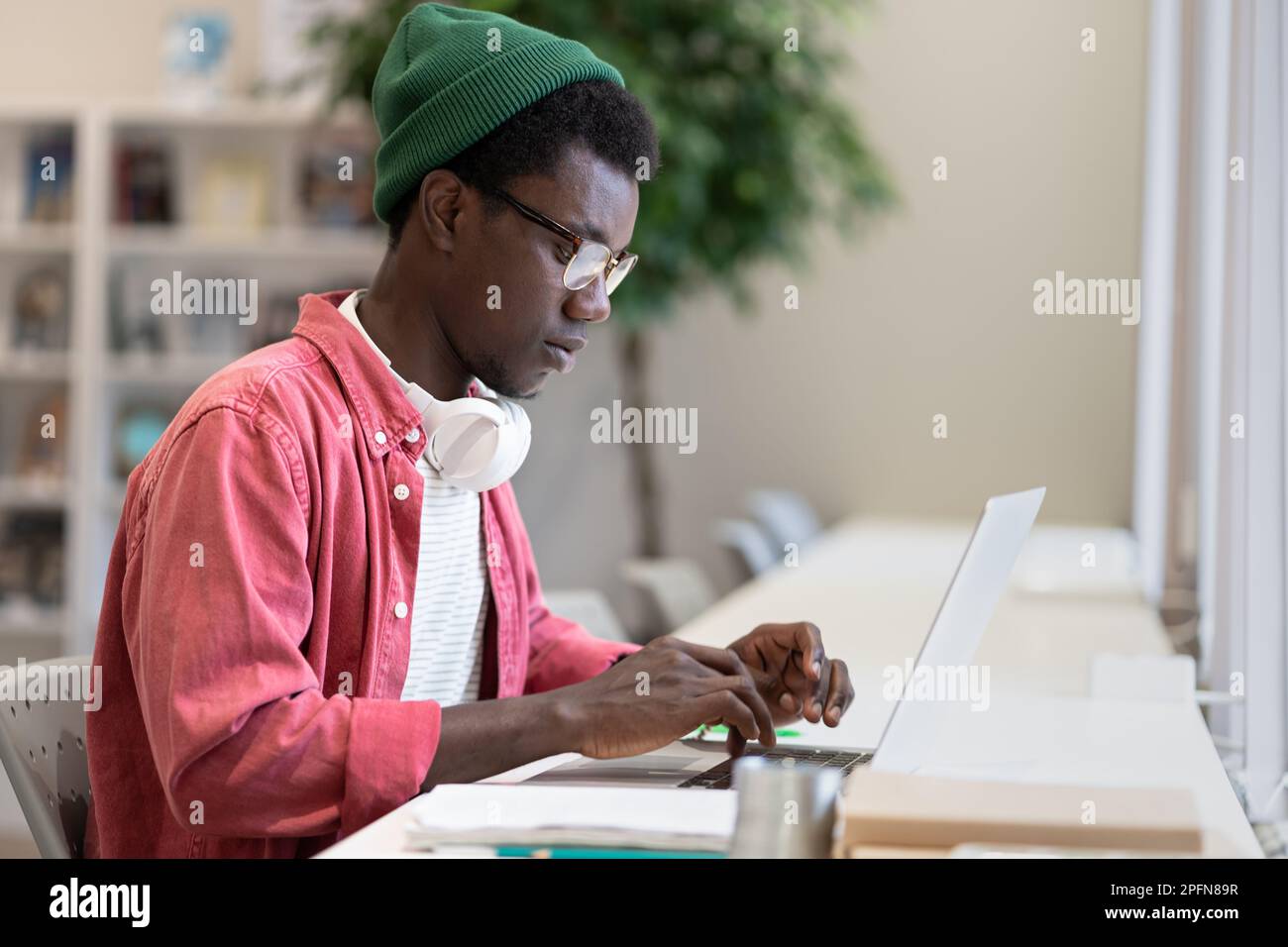 Junger afroamerikanischer Freiberufler, der an einem Laptop arbeitet und eine öffentliche Bibliothek als Arbeitsplatz verwendet Stockfoto