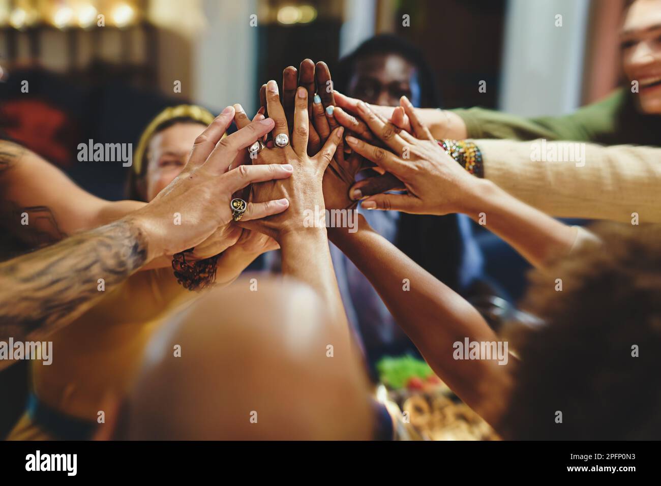 Eine vielfältige Gruppe von Freunden aus den Vierzigern mit verschiedenen ethnischen Zugehörigkeiten und Tattoos versammelt sich zum Abendessen an einem Tisch. Sie schließen sich enthusiastisch an den Händen Stockfoto