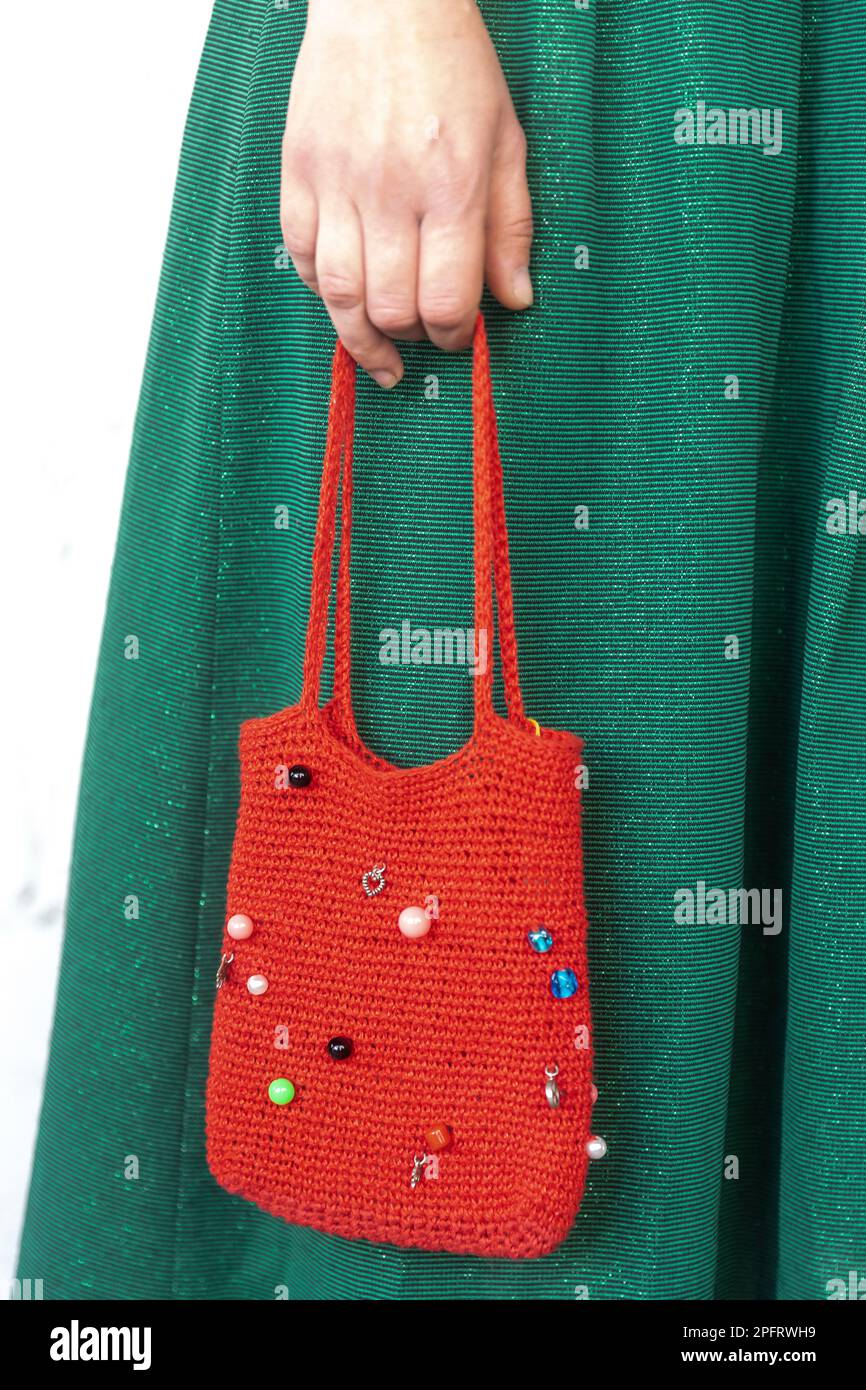 Eine rote Handtasche mit Strasssteinen in weiblichen Händen vor dem Hintergrund eines grünen Kleides. Stockfoto