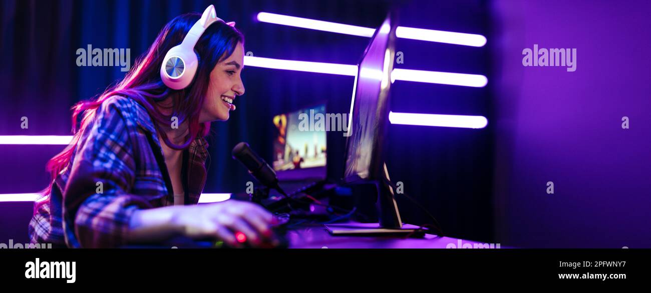Glückliche junge Frau streamt live ein Videospiel, sitzt vor einem Multi-Monitor-Setup mit einem Mikrofon für Kommentare und einem Headset für Kommunikation Stockfoto