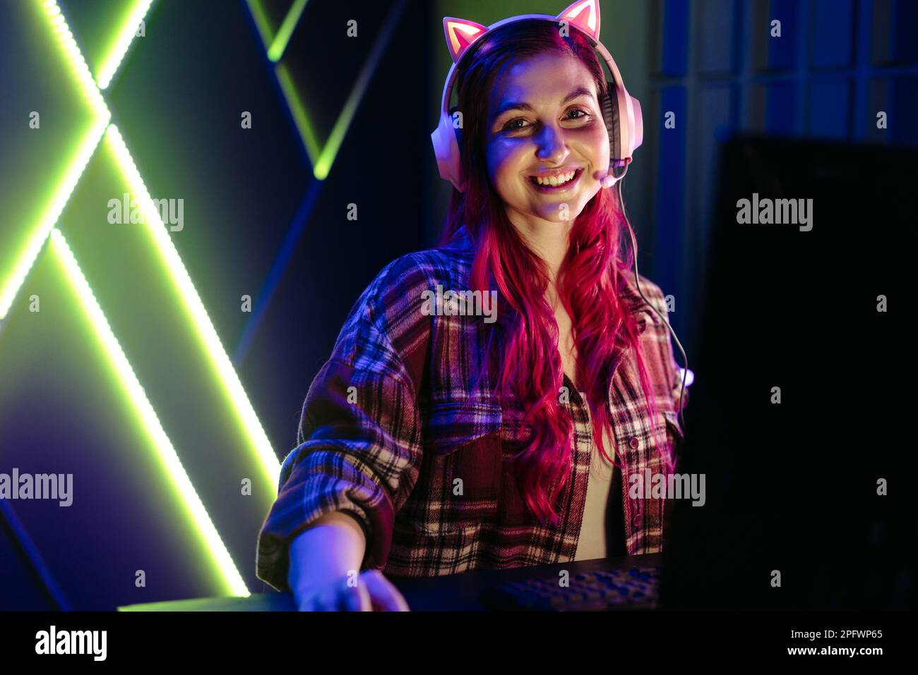 Porträt einer glücklichen jungen Frau, die mit einem Headset vor ihrem Gaming-pc sitzt. Eine Frau in ihren 20ern lächelt in die Kamera, während sie mit dem interagiert Stockfoto
