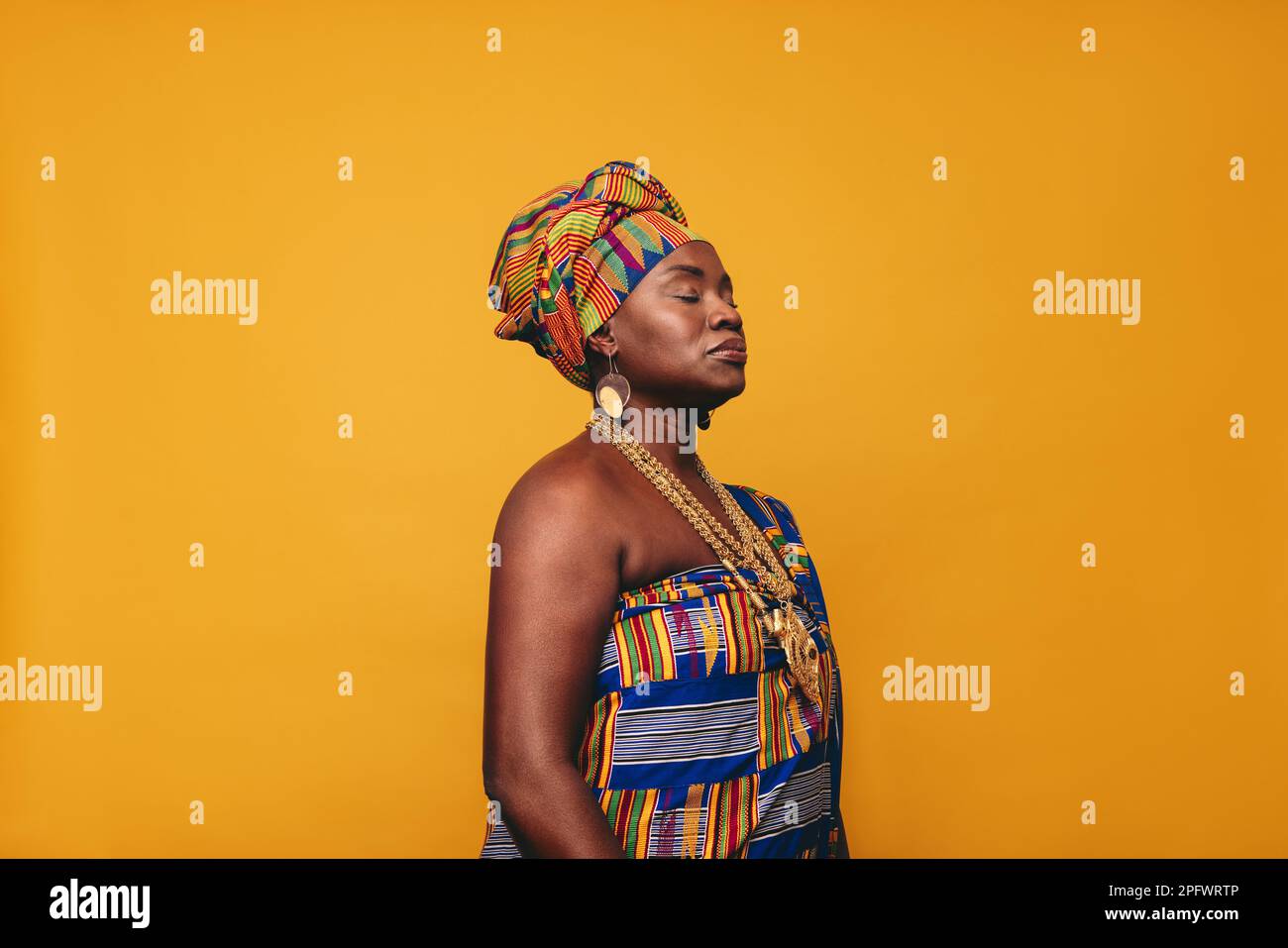 Reife Frau in traditioneller afrikanischer Kleidung auf gelbem Hintergrund. Selbstbewusste schwarze Frau in farbenfrohem Kente-Stoff und goldener Jewe Stockfoto