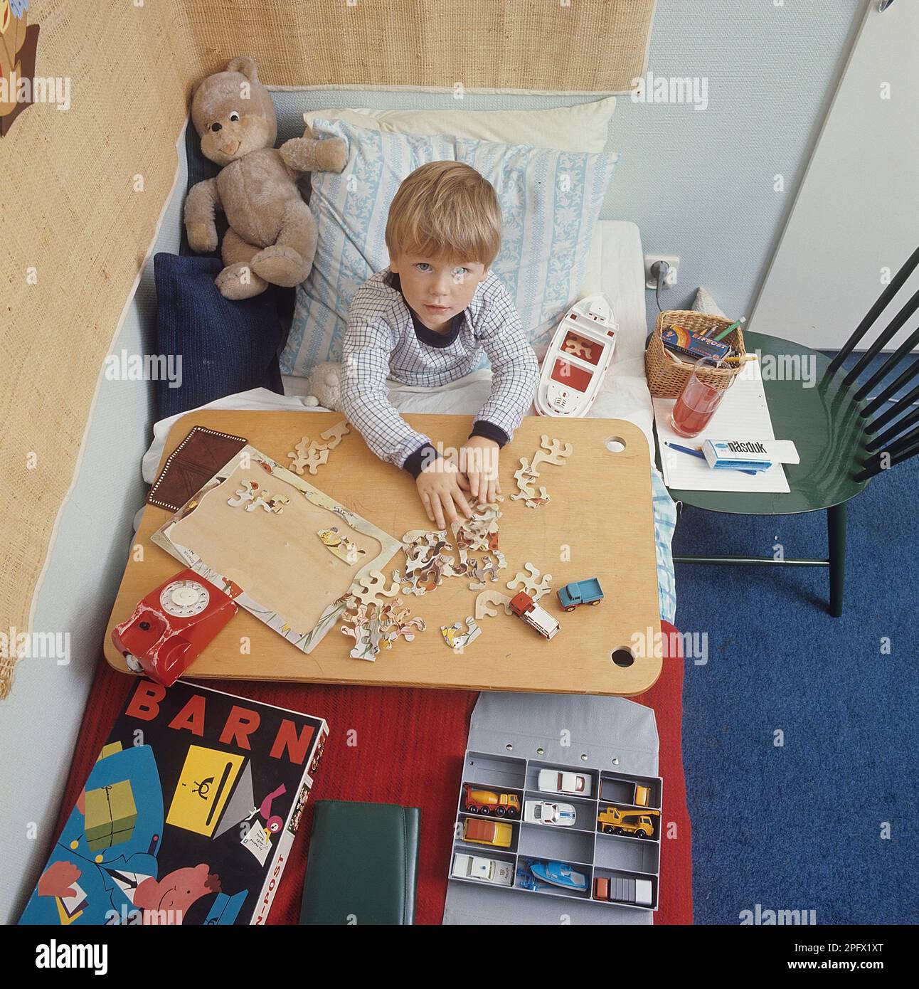 Ein Junge im Bett in den 1960er. Er hat wahrscheinlich eine Erkältung und muss zu Hause bleiben. Aber es besteht kein großer Bedarf für ihn. Er hat seine Spielzeuge wie Spielzeugautos, Boote und Stofftiere immer griffbereit. Auf dem Bild stellt er Rätsel zusammen. Schweden 1969 Stockfoto