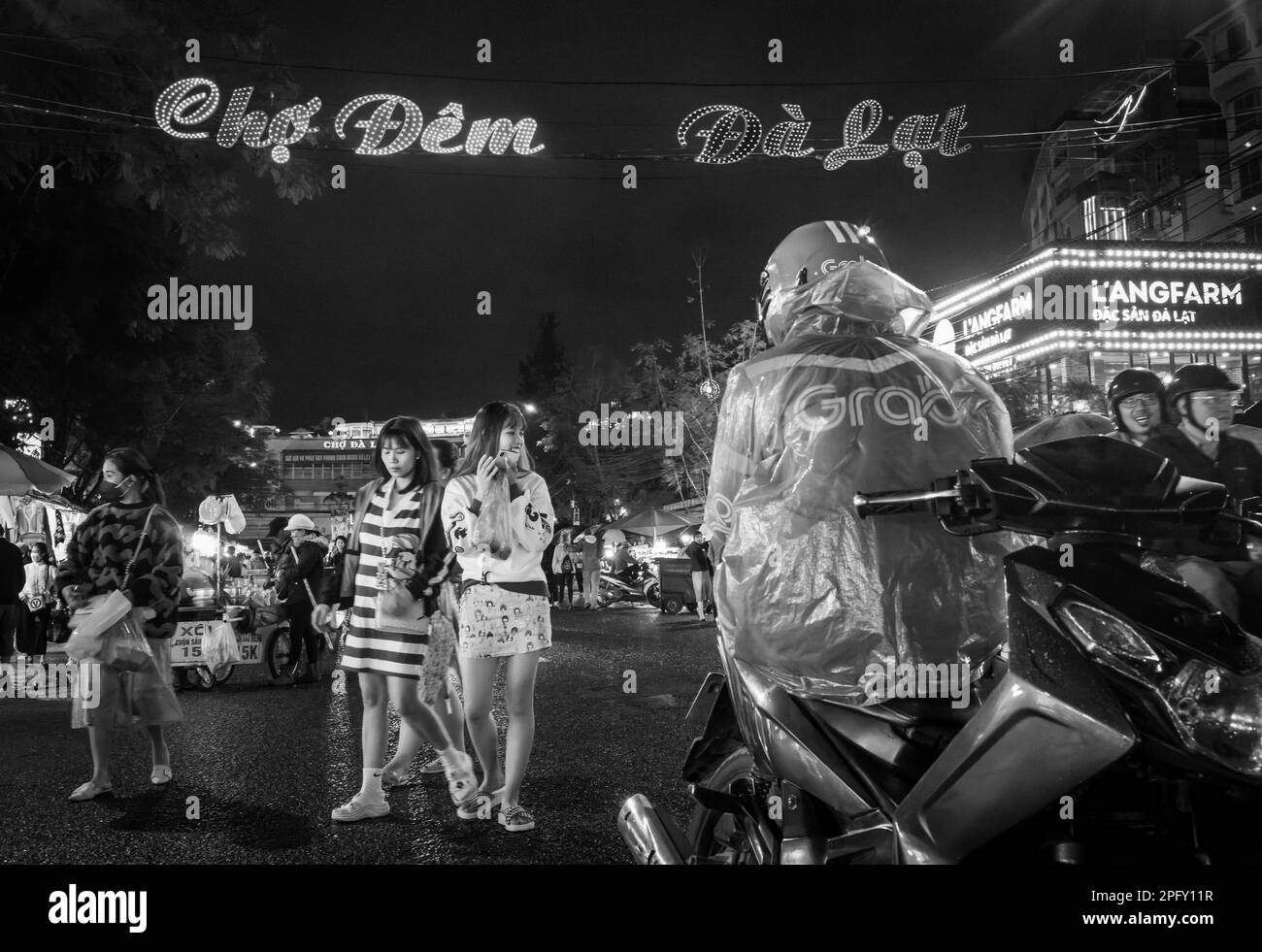 Zwei junge vietnamesische Mädchen gehen in Richtung eines Taxifahrers mit dem Motorrad, inmitten des geschäftigen Nachtmarkts in Dalat, Vietnam. Stockfoto