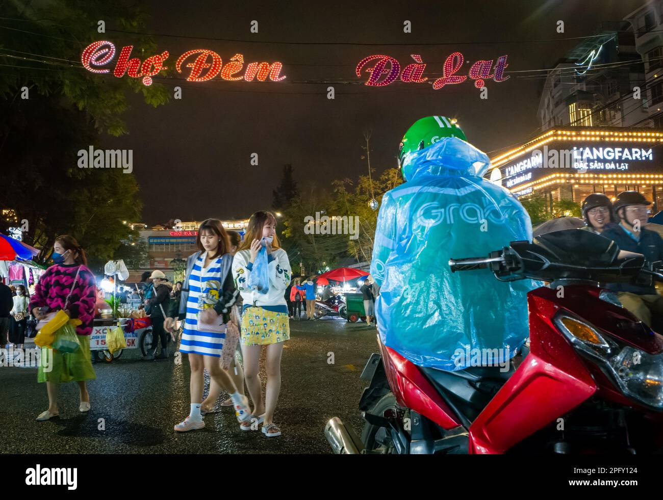 Zwei junge vietnamesische Mädchen gehen in Richtung eines Taxifahrers mit dem Motorrad, inmitten des geschäftigen Nachtmarkts in Dalat, Vietnam. Stockfoto