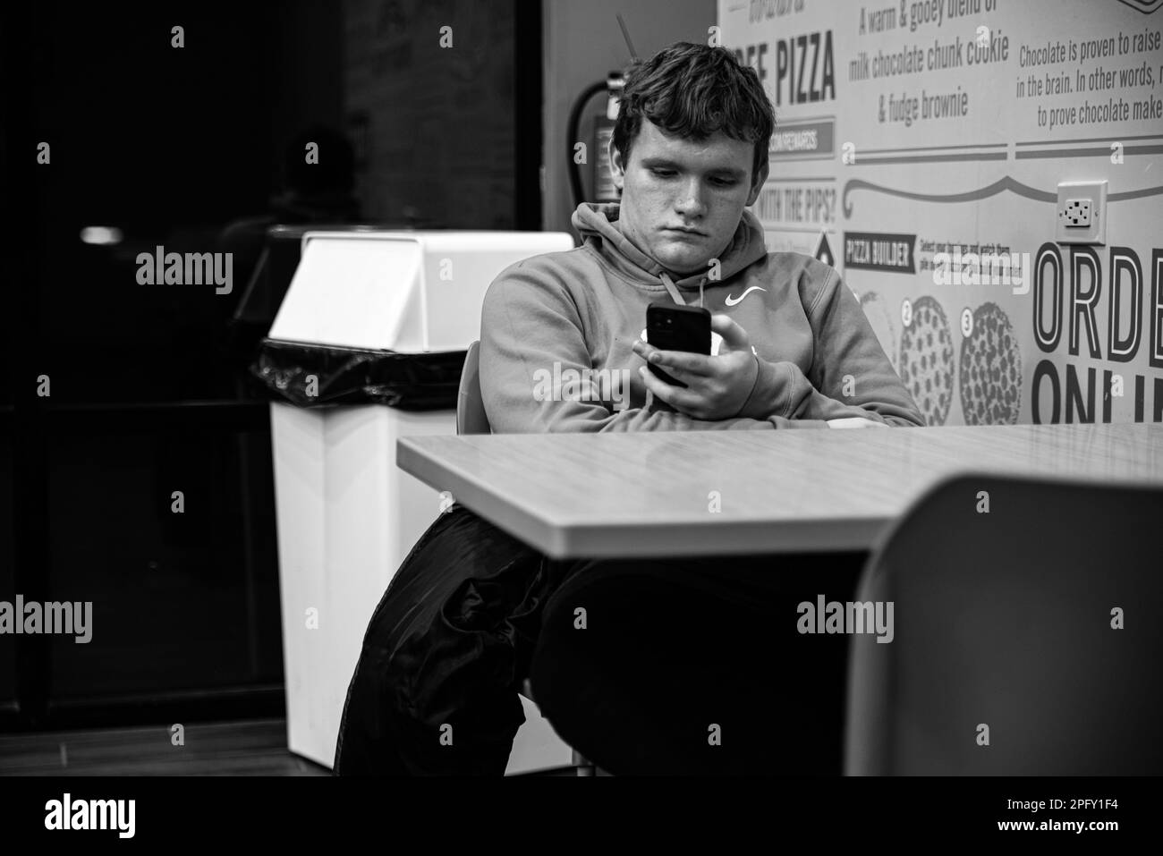 Ein junger weißer Mann sitzt an einem Tisch im Restaurant und sieht sich ein mobiles Gerät in seinen Händen an Stockfoto