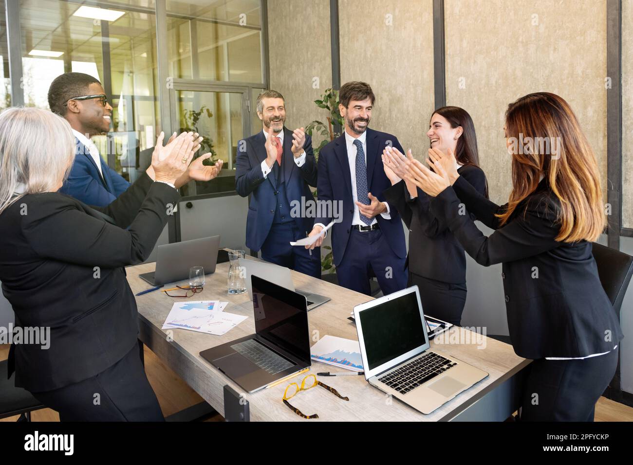 Ein Team von Geschäftsleuten verschiedener Rassen, die elegante Anzüge tragen, versammeln sich mit Laptops im Konferenzraum und applaudieren nach einem erfolgreichen Meeting Stockfoto