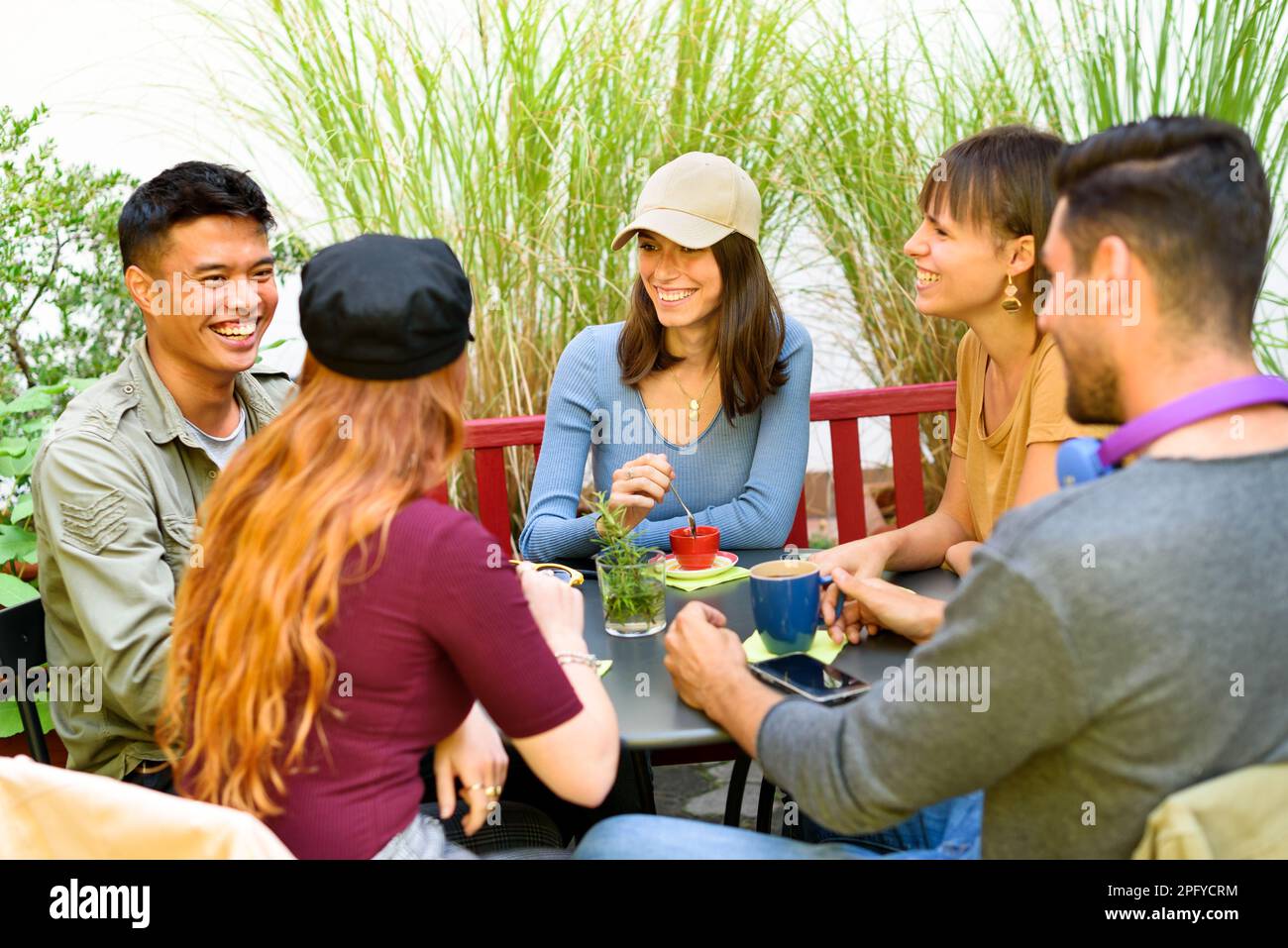 Eine Gruppe von fröhlichen, multiethnischen Freunden, die sich anschauen, während sie gemeinsam am Tisch frühstücken und Kaffee auf der Terrasse der Cafeteria im Freien trinken Stockfoto