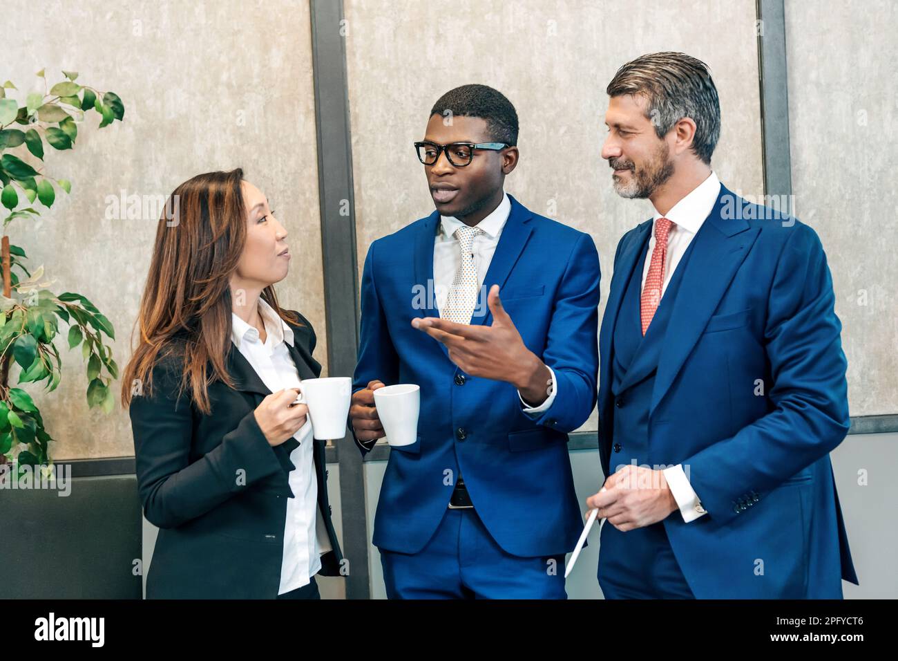 Multiethnische Kollegen in stilvollen Kleidern stehen mit Tassen heißer Getränke und sprechen während einer Pause von der Arbeit Stockfoto